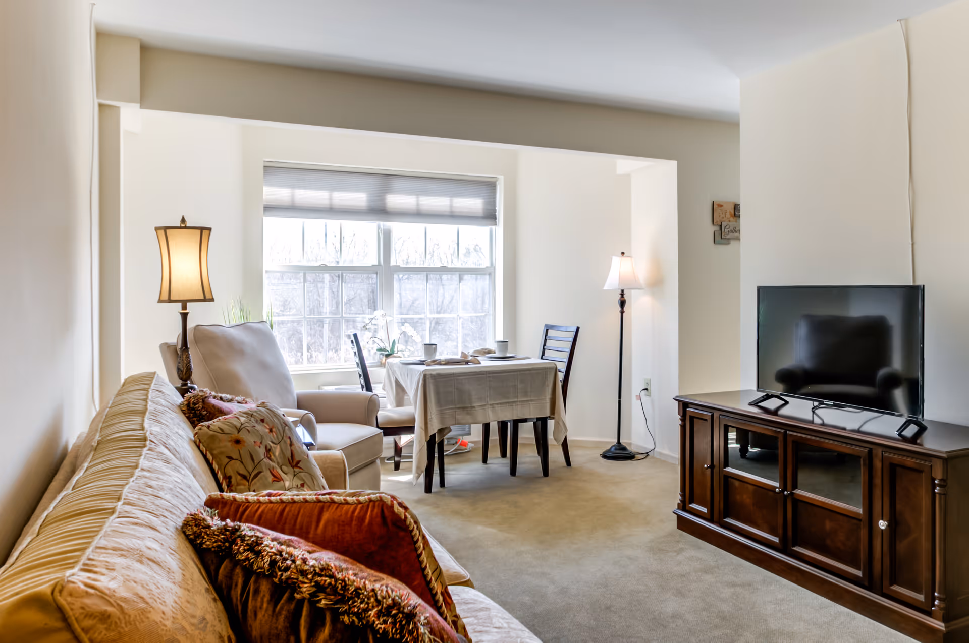 A cozy living room and dining area with a beige sofa adorned with decorative pillows, a cushioned armchair, a wooden TV stand with a flat-screen television, a small dining table covered with a white tablecloth, two chairs, and two standing lamps near a large window letting in natural light.