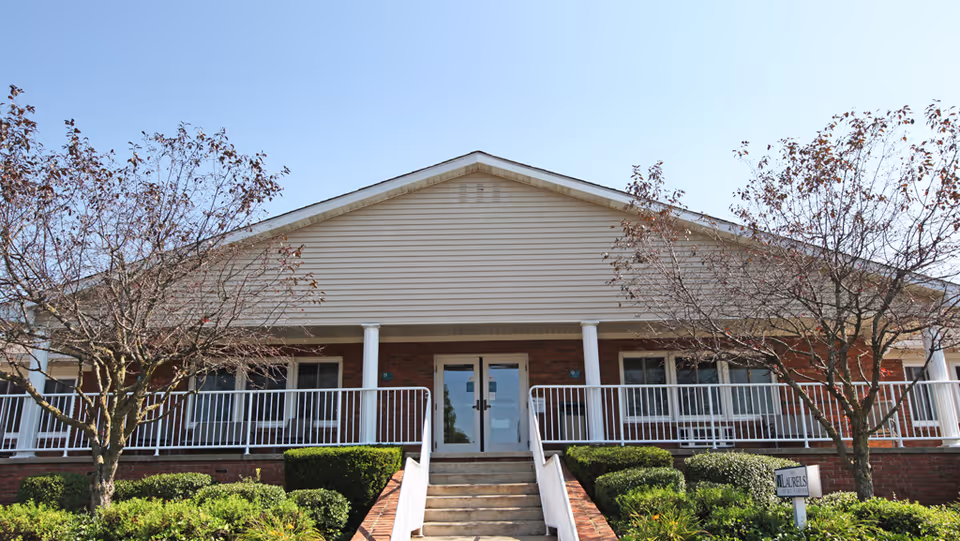 Front exterior view of a single-story building with a peaked roof, brick and siding facade, white railings, and a central staircase leading to double glass doors. There are two leafless trees and trimmed bushes in front of the building under a clear blue sky.