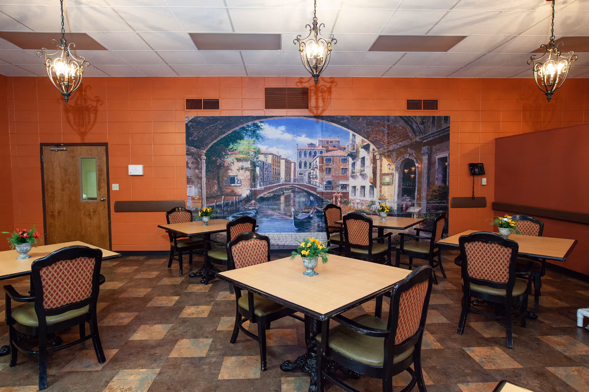 Dining area with several square tables and cushioned chairs arranged neatly. Each table has a small flower arrangement. The walls are painted orange, and there is a large mural depicting a Venetian canal scene with gondolas and historic buildings. Three decorative hanging light fixtures illuminate the room.