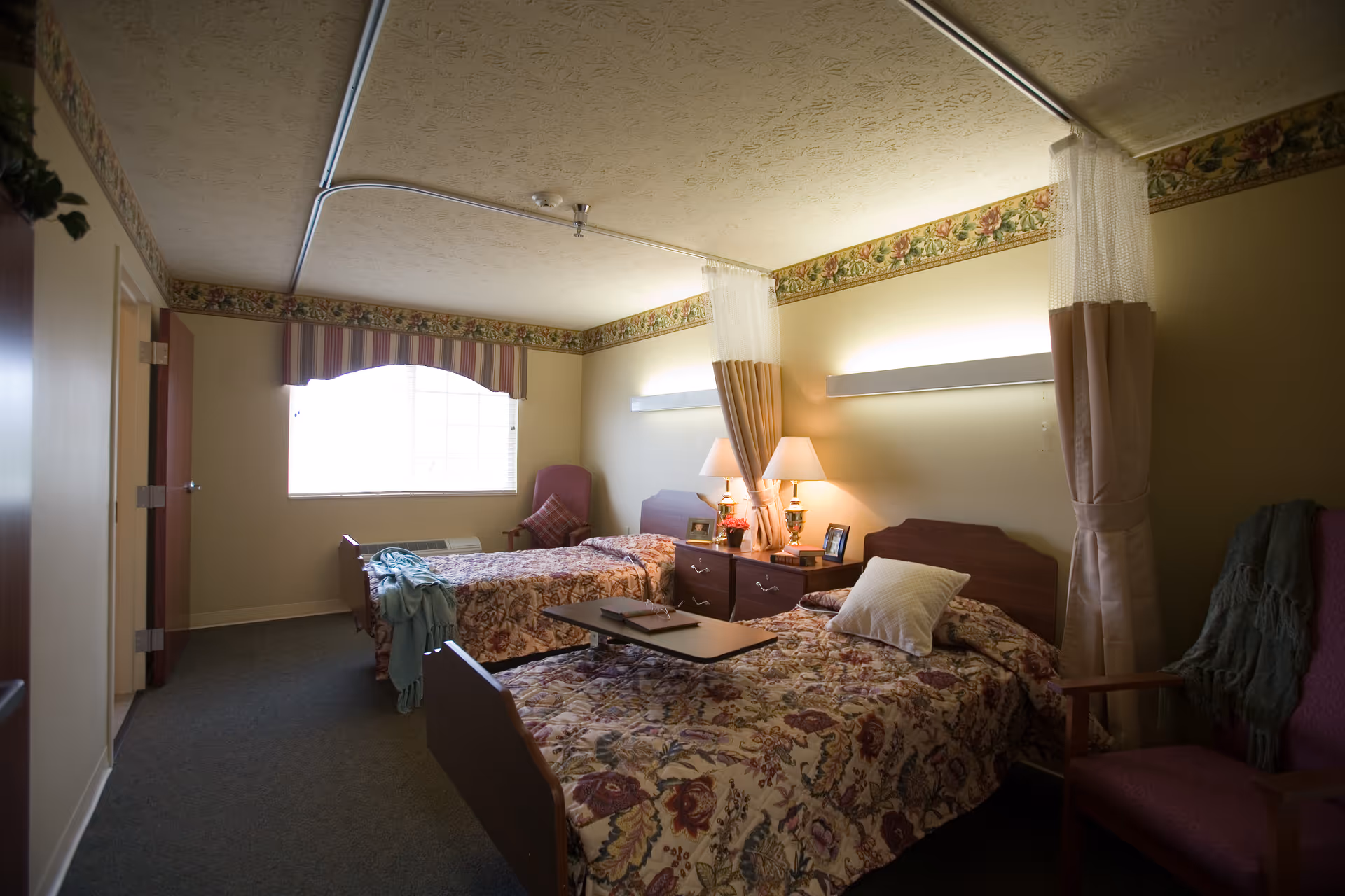 A shared senior living bedroom with two twin beds covered in floral bedspreads, a nightstand with lamps between them, privacy curtains, and a window with a valance.