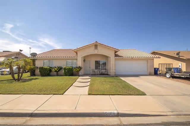 Single-story house with a tiled roof, a small front porch with a table and chairs, a two-car garage, and a well-maintained lawn with some bushes and palm trees under a clear blue sky.