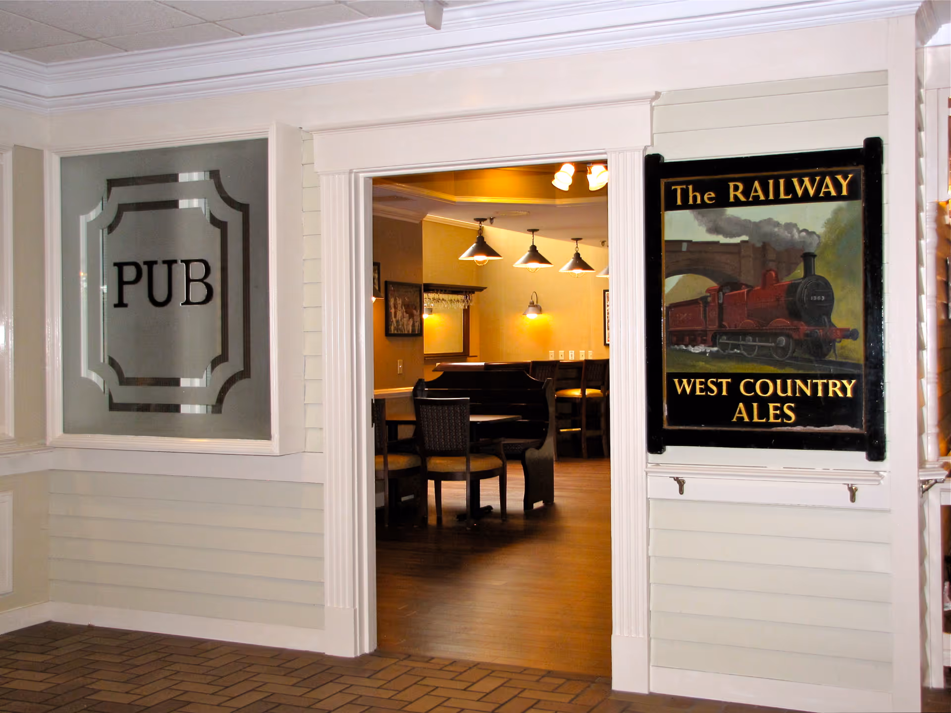 Entrance to a pub area inside a facility with a frosted glass window labeled 'PUB' on the left and a vintage-style sign on the right that reads 'The RAILWAY WEST COUNTRY ALES' featuring a red steam train. Inside, there are wooden tables and chairs with warm lighting fixtures hanging from the ceiling.