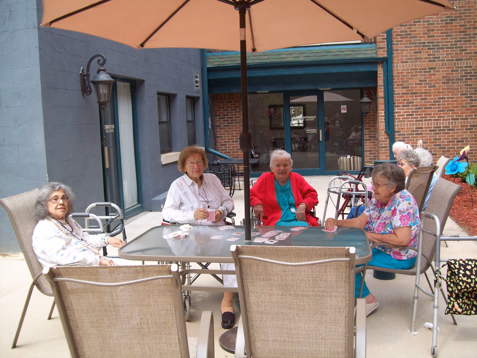 A group of elderly women sitting around a glass outdoor table under a large umbrella, playing cards. They are seated in patio chairs on a concrete surface outside a brick and blue-painted building. Some walkers are visible near the women, and there are plants and mulch along the building edge.