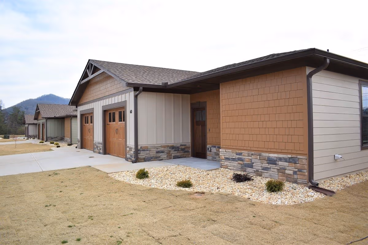 Exterior view of a single-story residential building with a two-car garage, stone and wood siding, and a small landscaped area with rocks and shrubs in front. The building is part of a row of similar houses with mountains visible in the background.