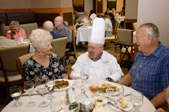 A chef wearing a white uniform and tall chef hat is seated at a dining table with an elderly woman and man in a senior living community dining room. Plates of food and glasses of water and wine are on the table. Other elderly residents are visible dining in the background.
