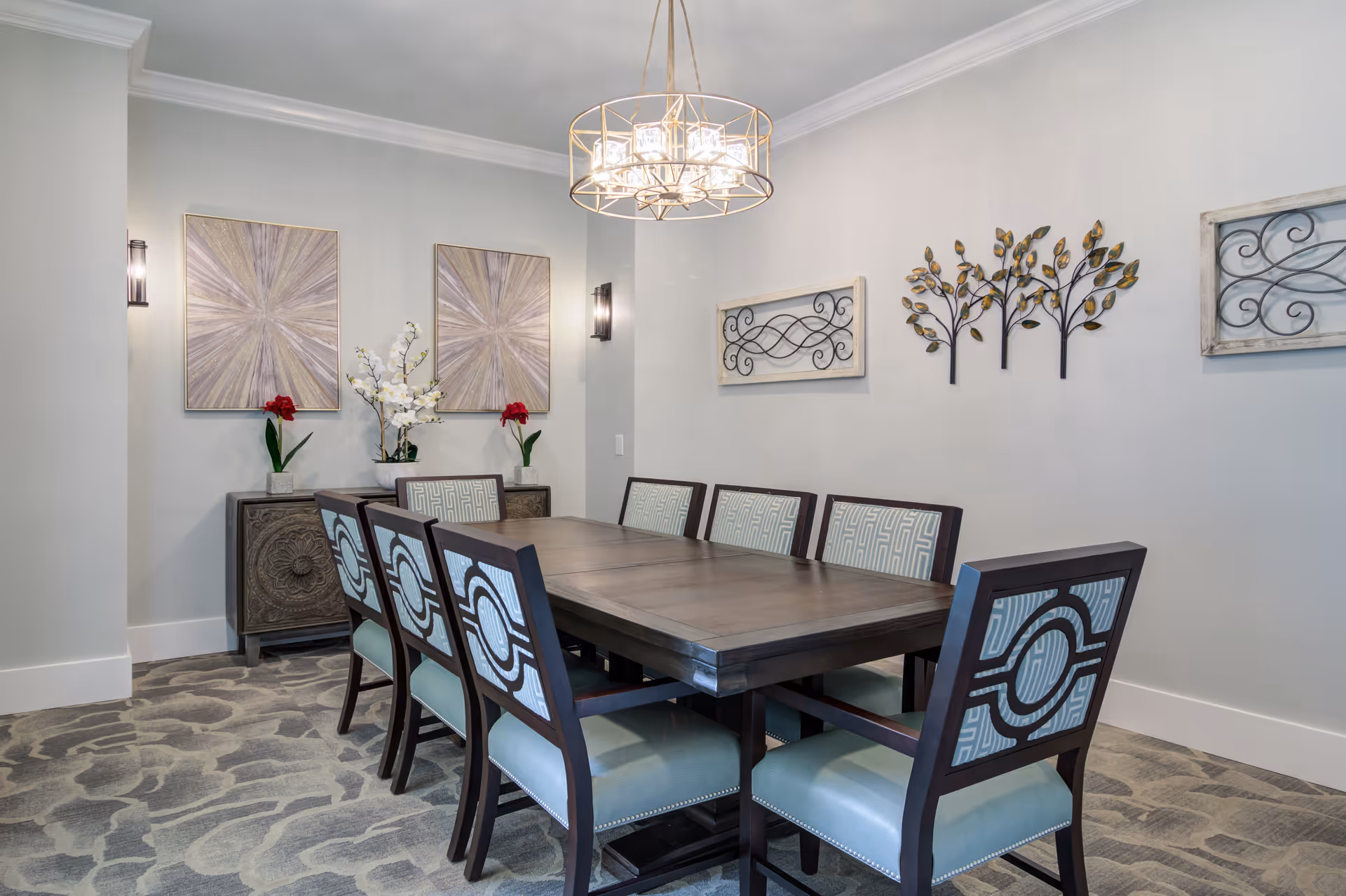 A modern dining room with a large dark wooden table surrounded by eight upholstered chairs with patterned backs and light blue seats. The walls are light gray and decorated with two abstract paintings, metal wall art shaped like trees, and two framed scroll designs. A contemporary chandelier with multiple lights hangs above the table, and a sideboard with flower arrangements is positioned against the wall.