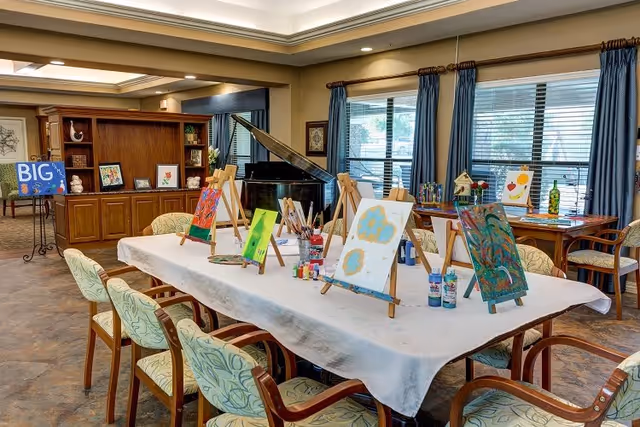 A well-lit activity room with a long table covered by a white cloth, set up for painting with multiple easels holding colorful paintings, paint bottles, and brushes. The room has several chairs around the table, large windows with blue curtains, a grand piano in the background, and wooden shelves with decorative items.