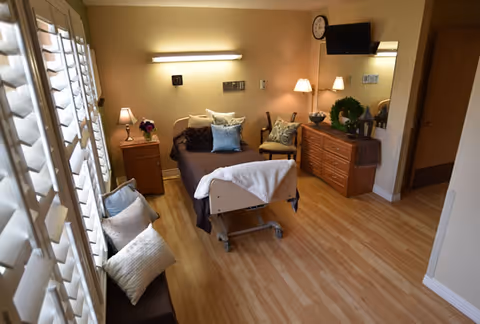 A well-lit room with a single hospital-style bed covered with brown bedding and multiple pillows. The room has wooden flooring, a wooden dresser with decorative items including a green wreath, a chair with a cushion, a wall-mounted TV, and a clock. There are large windows with white shutters on the left side, and a bench with several pillows beneath the windows.