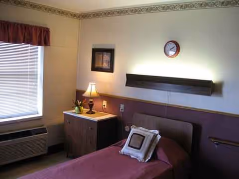 A single bedroom with purple bedspread, bedside cabinet, lamp, window blinds, and wall clock.