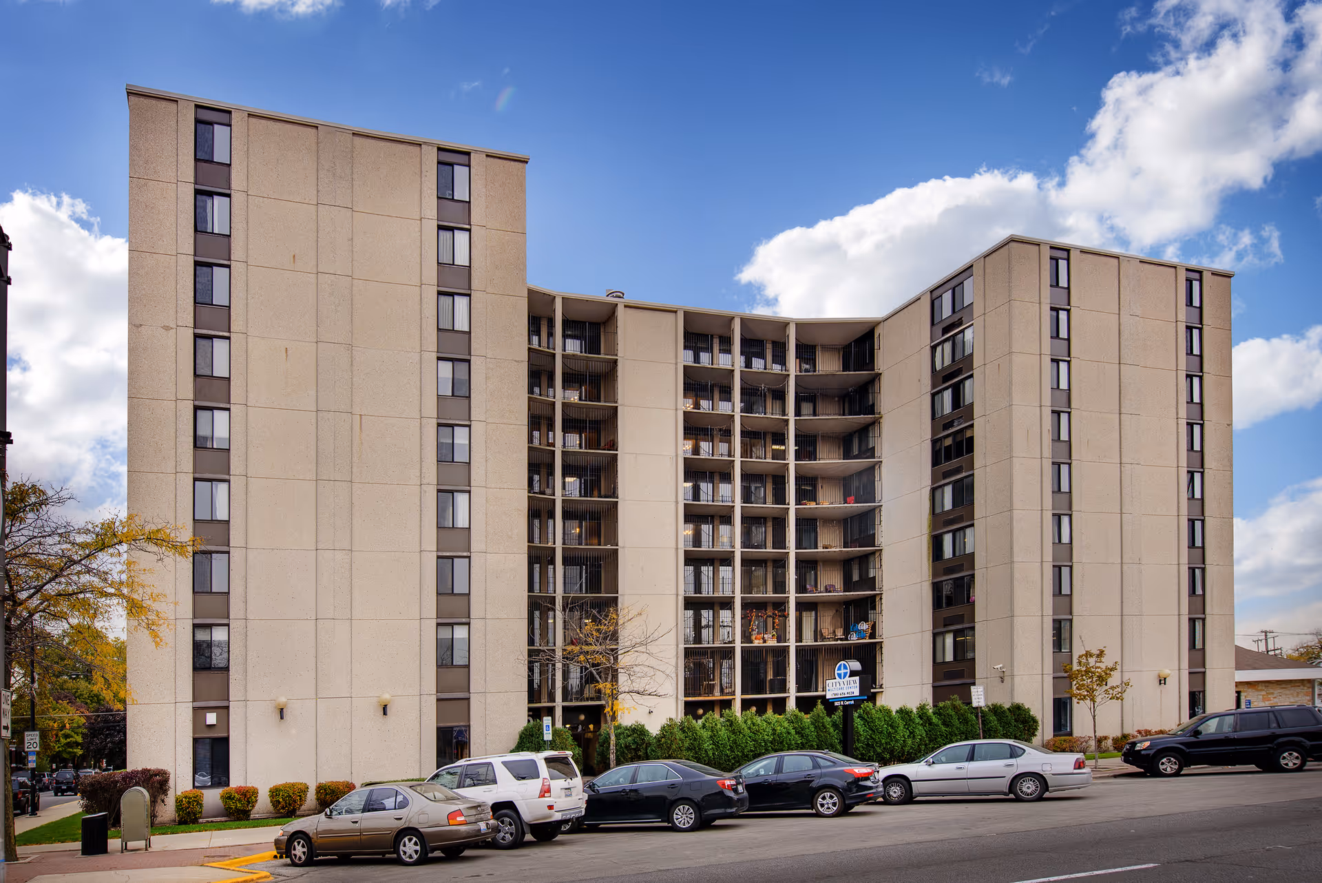 Exterior view of a multi-story senior living facility named City View Multicare Center with parked cars along the street in front and a clear blue sky with some clouds.