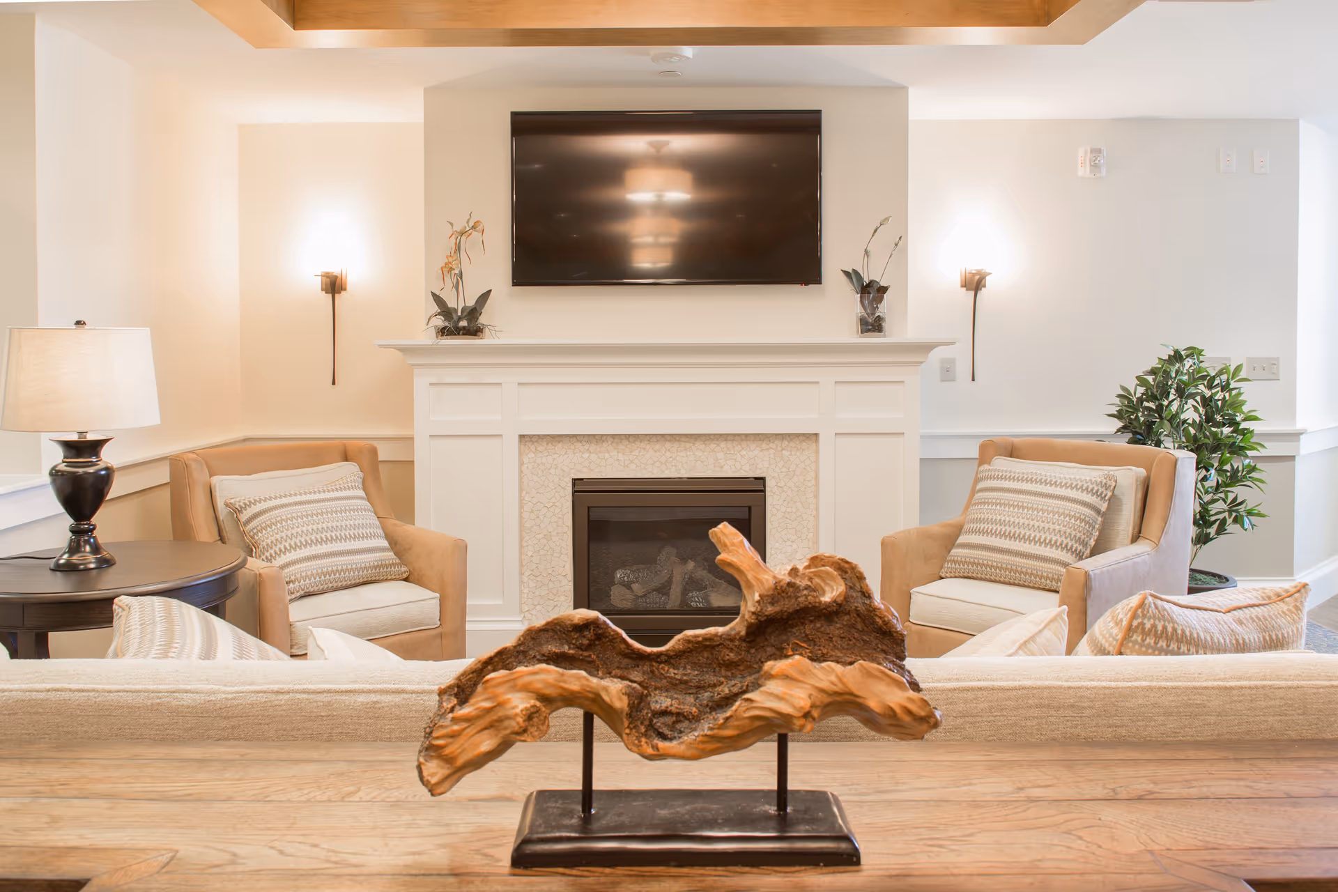 A cozy living room area featuring a white fireplace with a mounted flat-screen TV above it. Two beige armchairs with patterned cushions flank the fireplace, accompanied by a side table with a lamp on the left. A decorative wooden sculpture is placed on a wooden table in the foreground, and a green potted plant is visible on the right side of the room. The walls are painted white with soft lighting from wall sconces.