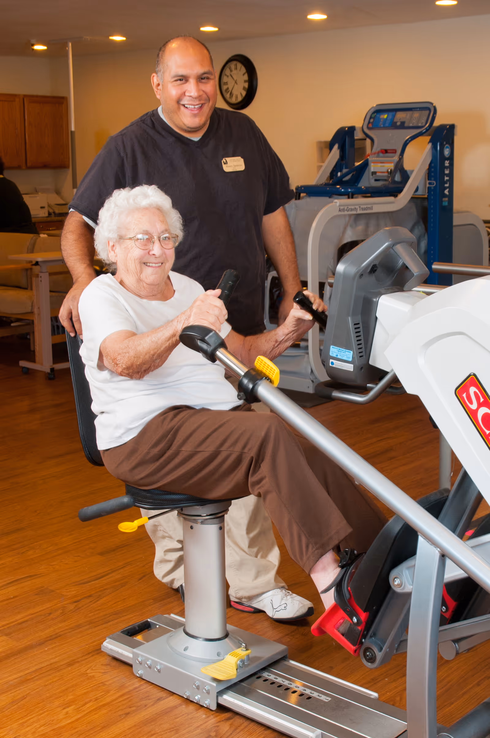 An elderly woman using a seated exercise machine in a fitness room, assisted by a smiling male staff member standing behind her. The room has wooden floors, exercise equipment, and a clock on the wall.