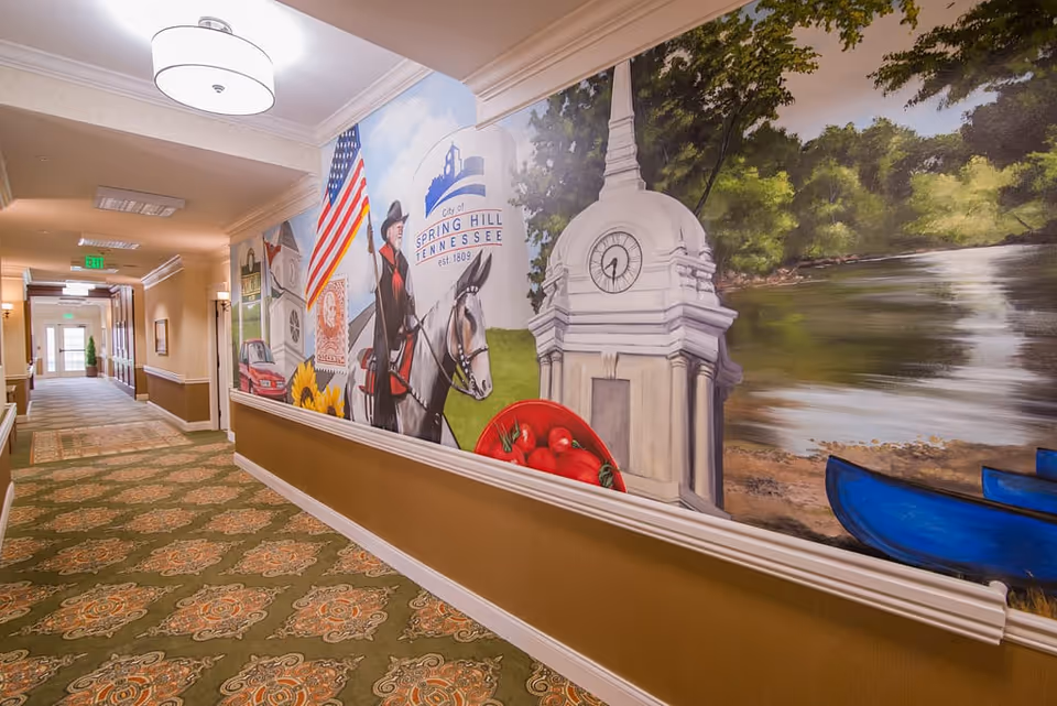 Interior hallway of Morning Pointe of Spring Hill featuring a mural on the right wall depicting various local scenes including a man on horseback, an American flag, a clock tower, tomatoes, and a river with a blue canoe. The hallway has patterned carpet, beige walls with white trim, and ceiling lights.