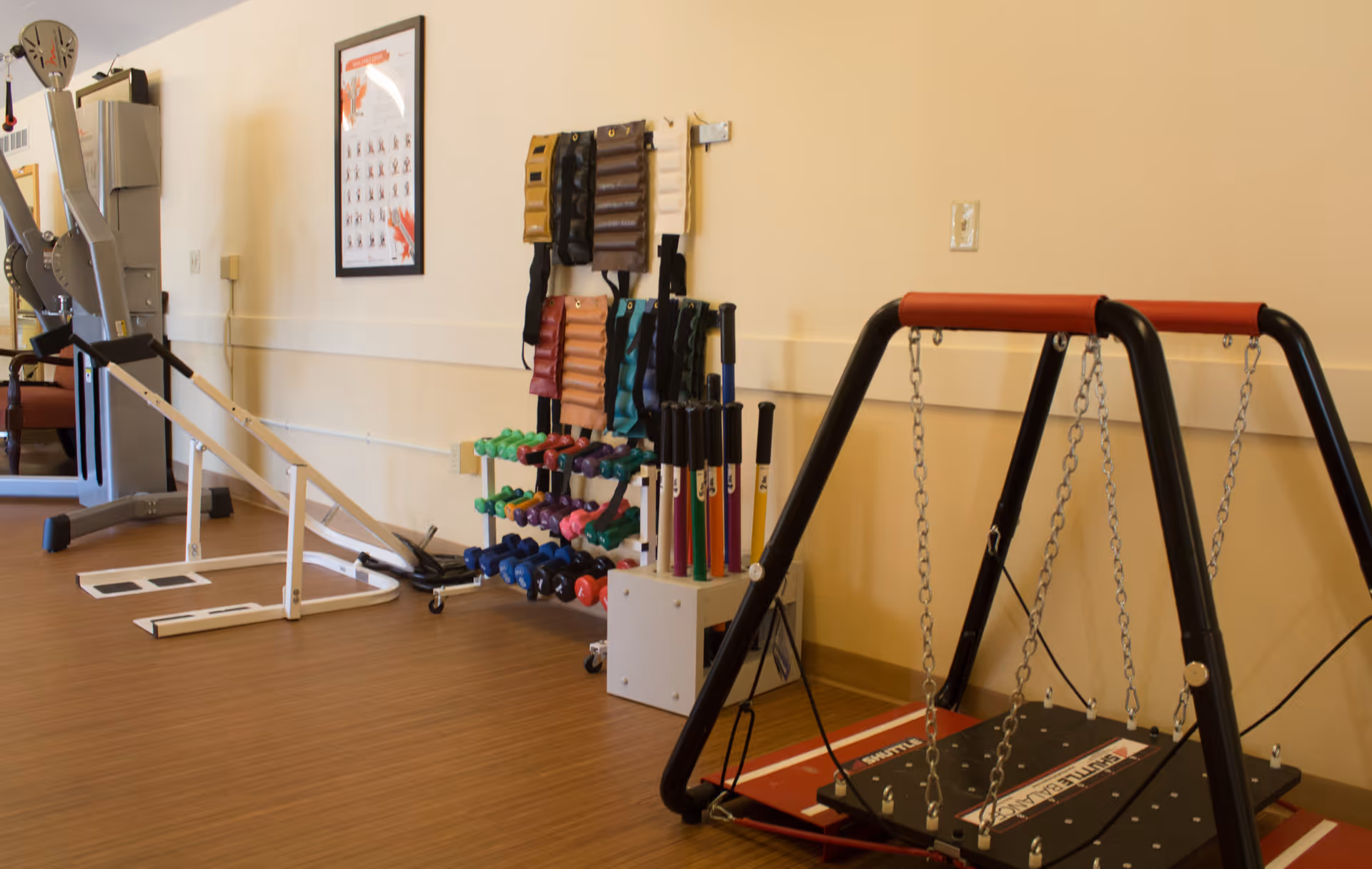 A senior living facility exercise room with various fitness equipment including a balance board, hand weights, resistance bands, and a black and red platform swing. The room has light-colored walls and wood flooring.