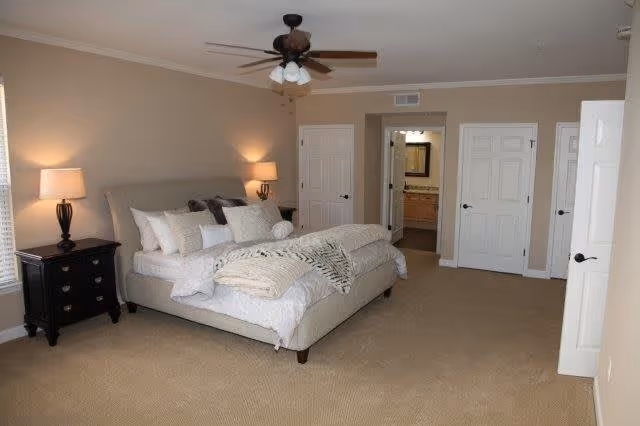 A spacious bedroom with a large bed featuring multiple pillows and a blanket. There are two bedside tables with lamps on each side of the bed. The room has beige walls and carpeted floor. A ceiling fan with lights is mounted on the ceiling. There are three white doors visible, one leading to a bathroom with a vanity and mirror.