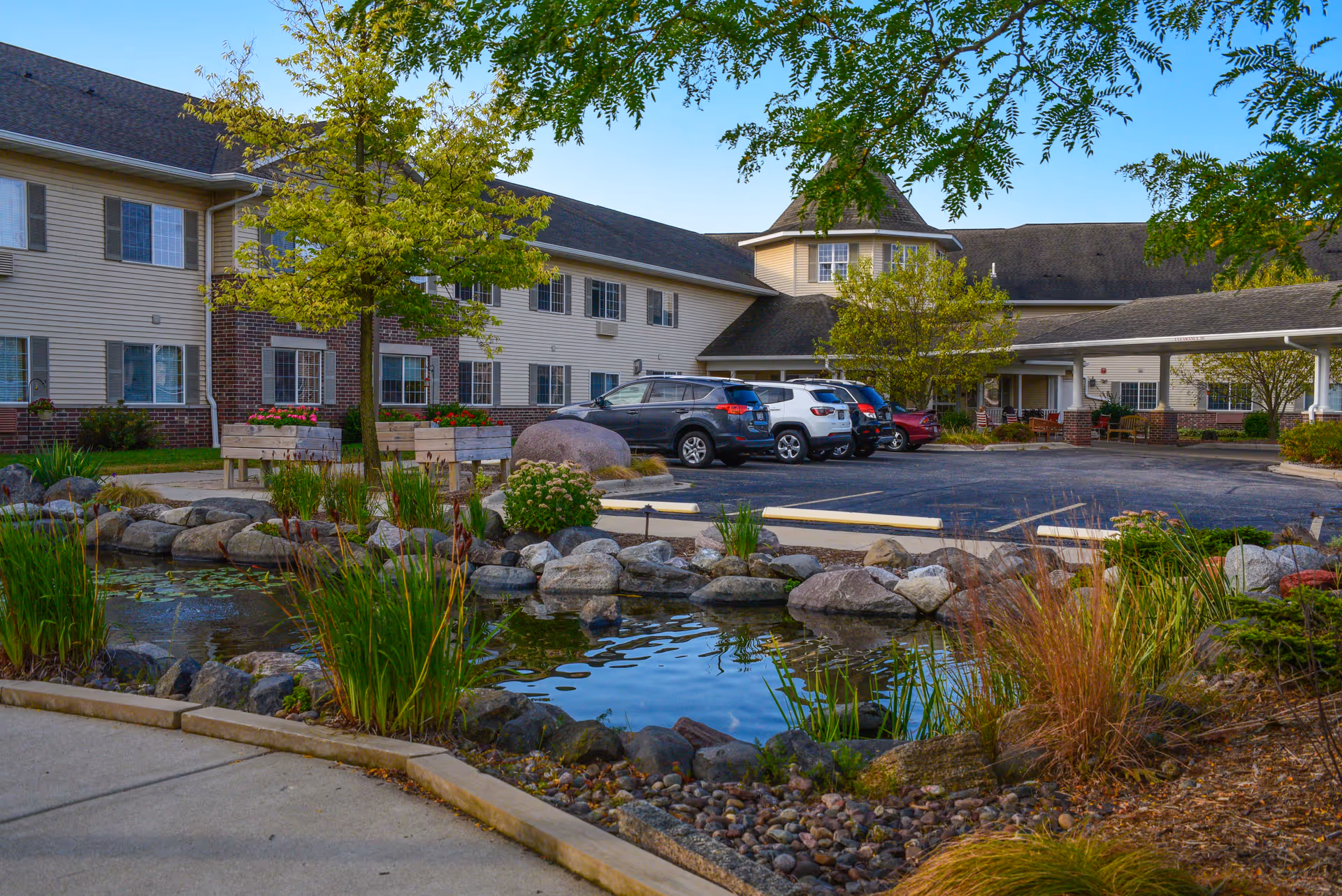Exterior view of Drumlin Reserve - Assisted Living facility showing a landscaped pond with rocks and plants in the foreground, a parking lot with several cars, and a two-story building with beige siding and brick accents in the background under a clear sky.