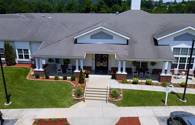 Front entrance of a single-story senior living building with a covered porch, rocking chairs, and landscaped walkway.