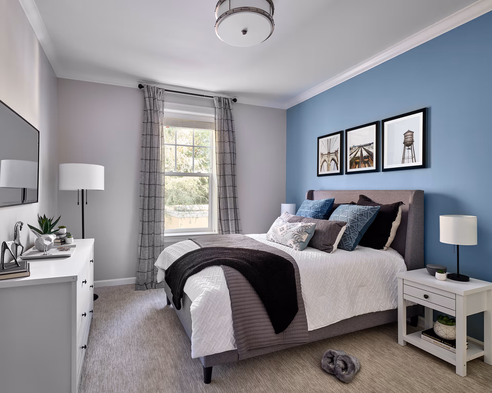 A neatly arranged bedroom with a bed featuring multiple pillows and a white textured bedspread with a dark throw blanket. The wall behind the bed is painted blue and decorated with three framed pictures. There is a white nightstand with a lamp and small plant on the right side of the bed, and a white dresser with decorative items and a floor lamp on the left side. A window with gray plaid curtains lets in natural light, and a ceiling light fixture is visible.