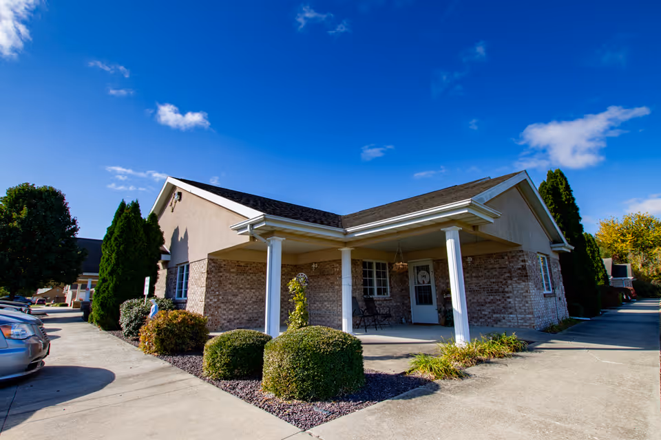 Exterior view of a single-story brick building with a covered porch supported by white columns, surrounded by neatly trimmed bushes and trees under a bright blue sky with a few clouds. There are parked cars along the driveway.
