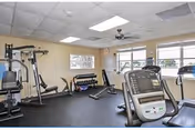 A fitness room with various exercise equipment including a multi-station gym machine, dumbbells on a rack, a bench, and a treadmill. The room has beige walls, large windows letting in natural light, a ceiling fan, and a black rubber floor.
