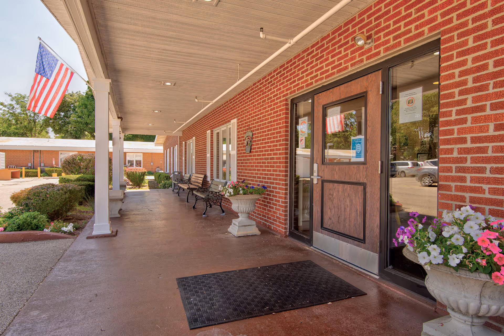 Covered brick entrance of a senior living facility with benches, potted flowers, an American flag, and a glass door.