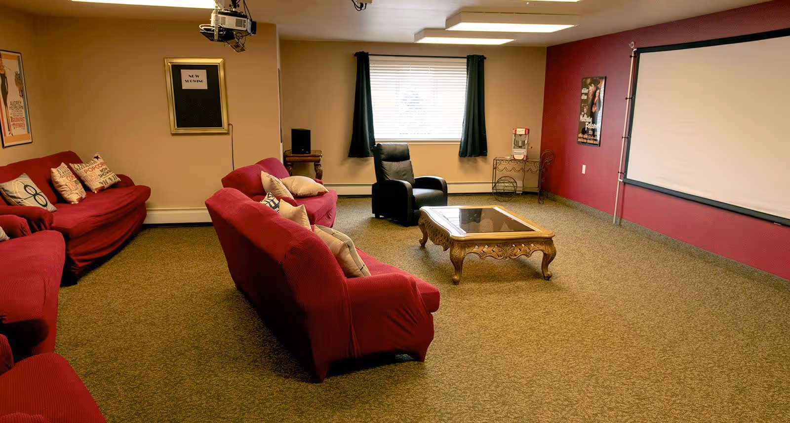 A cozy living room with red couches covered in red slipcovers and decorated with various pillows. There is a black recliner chair near a window with dark curtains. A wooden coffee table with a glass top sits in the center of the room. On the right wall, there is a large white projection screen and a movie poster. The walls are painted beige and red, and the floor is carpeted.
