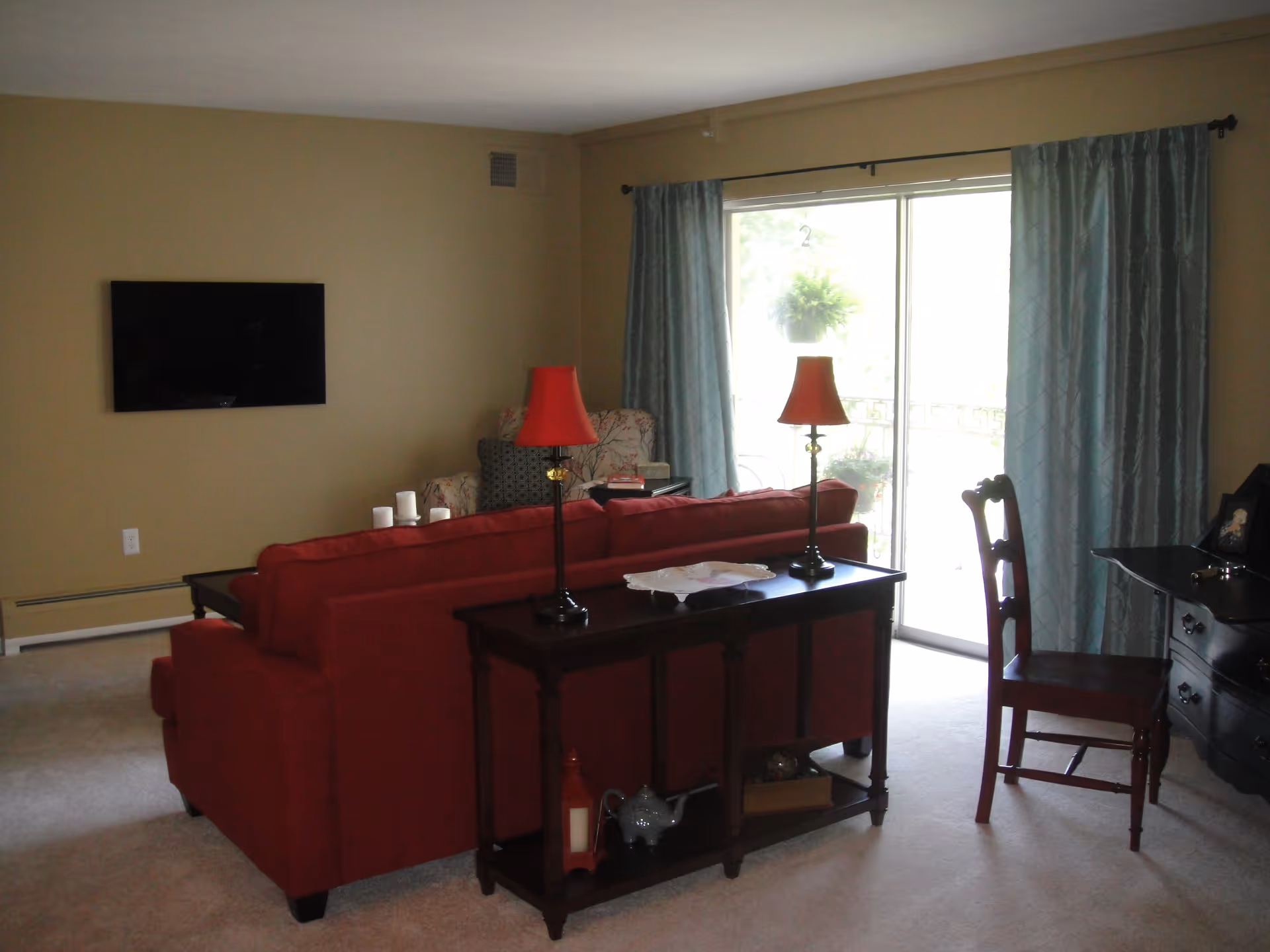 A living room with a red sofa facing a wall-mounted flat screen TV. Behind the sofa is a dark wooden console table with two red lamps and decorative items. To the right, there is a wooden chair and a dark wooden desk. Light blue curtains frame a sliding glass door leading to an outdoor area.