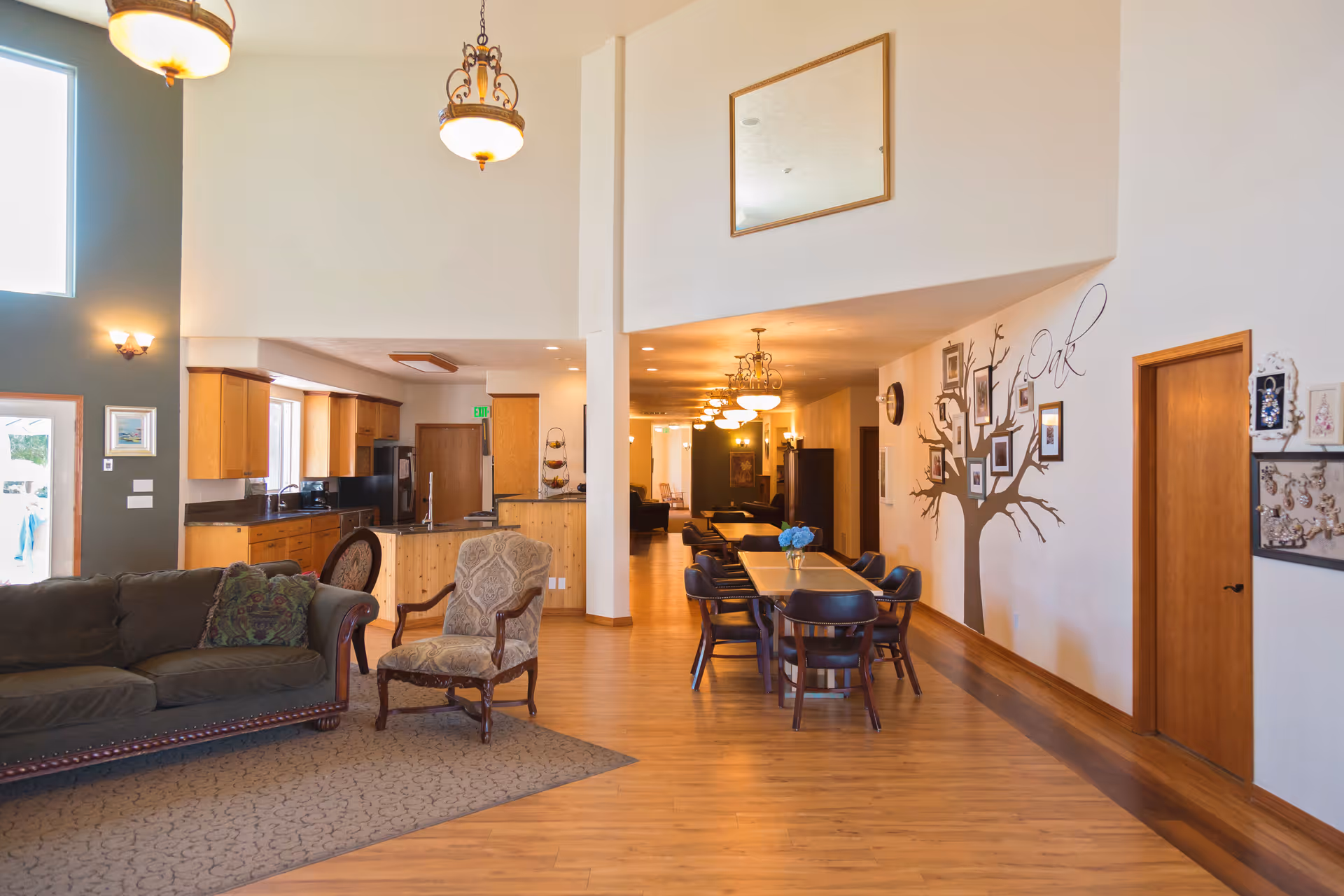 Spacious interior of a senior living facility featuring a living area with a dark green sofa and an upholstered armchair on a carpet, an open kitchen with wooden cabinets and black appliances, and a dining area with a long wooden table and several chairs. The walls are decorated with framed pictures and a tree mural with the word 'Oak'. The space has wooden flooring, high ceilings, and multiple hanging light fixtures.