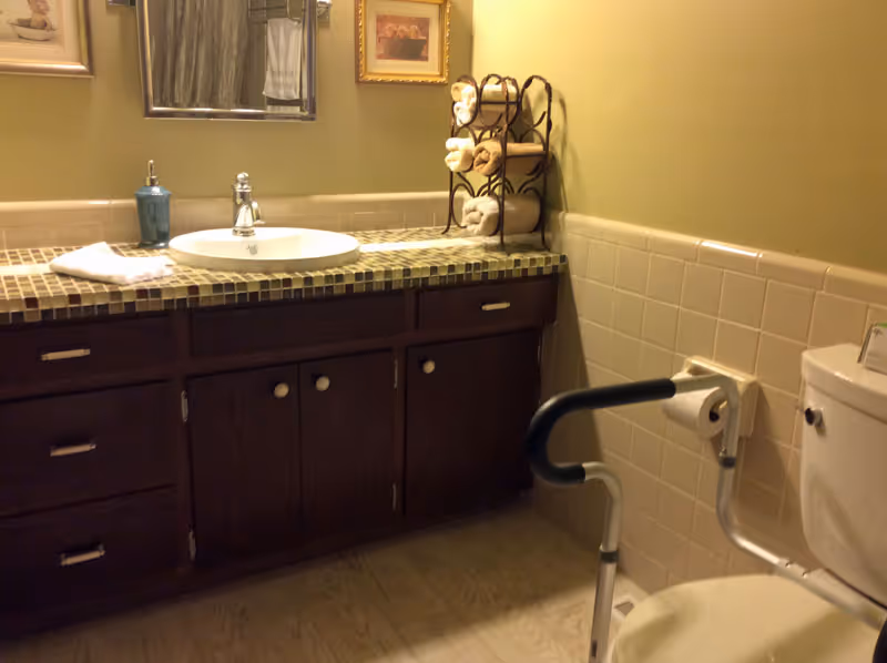 Bathroom with a tiled vanity and sink, mirror and towel rack, and a toilet equipped with a safety grab bar.