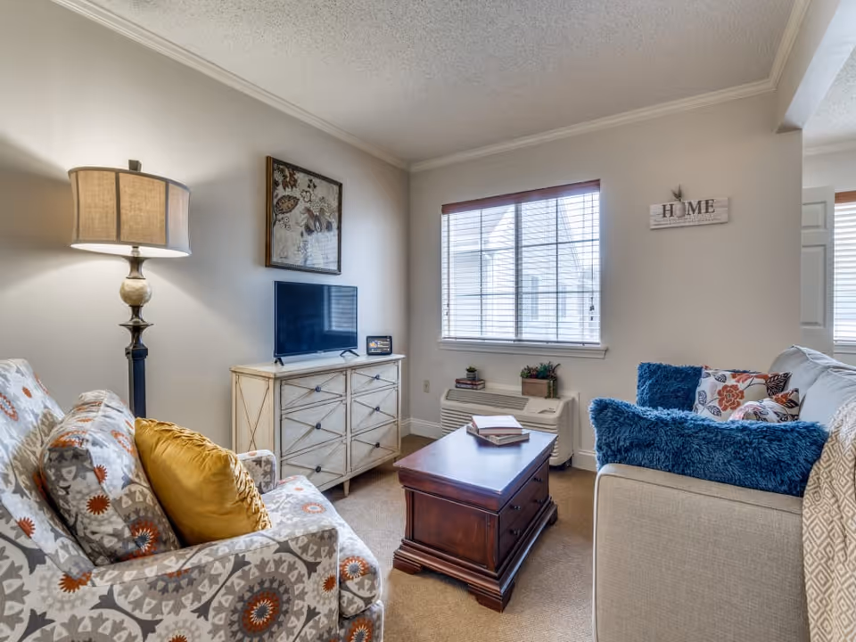 A cozy living room with a patterned armchair and a beige sofa adorned with blue and floral pillows. A wooden coffee table with books sits in the center. A floor lamp stands beside the armchair, and a TV is placed on a white dresser against the wall. A window with blinds lets in natural light, and a decorative sign that reads 'HOME' is mounted on the wall.