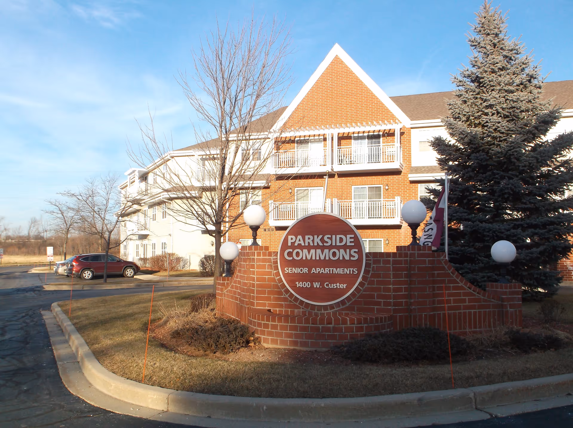 Exterior view of Parkside Commons Senior Apartments showing a brick sign with the facility name and address 1400 W. Custer, surrounded by a landscaped area with grass, bushes, and trees. The building is visible in the background with balconies and a clear blue sky.