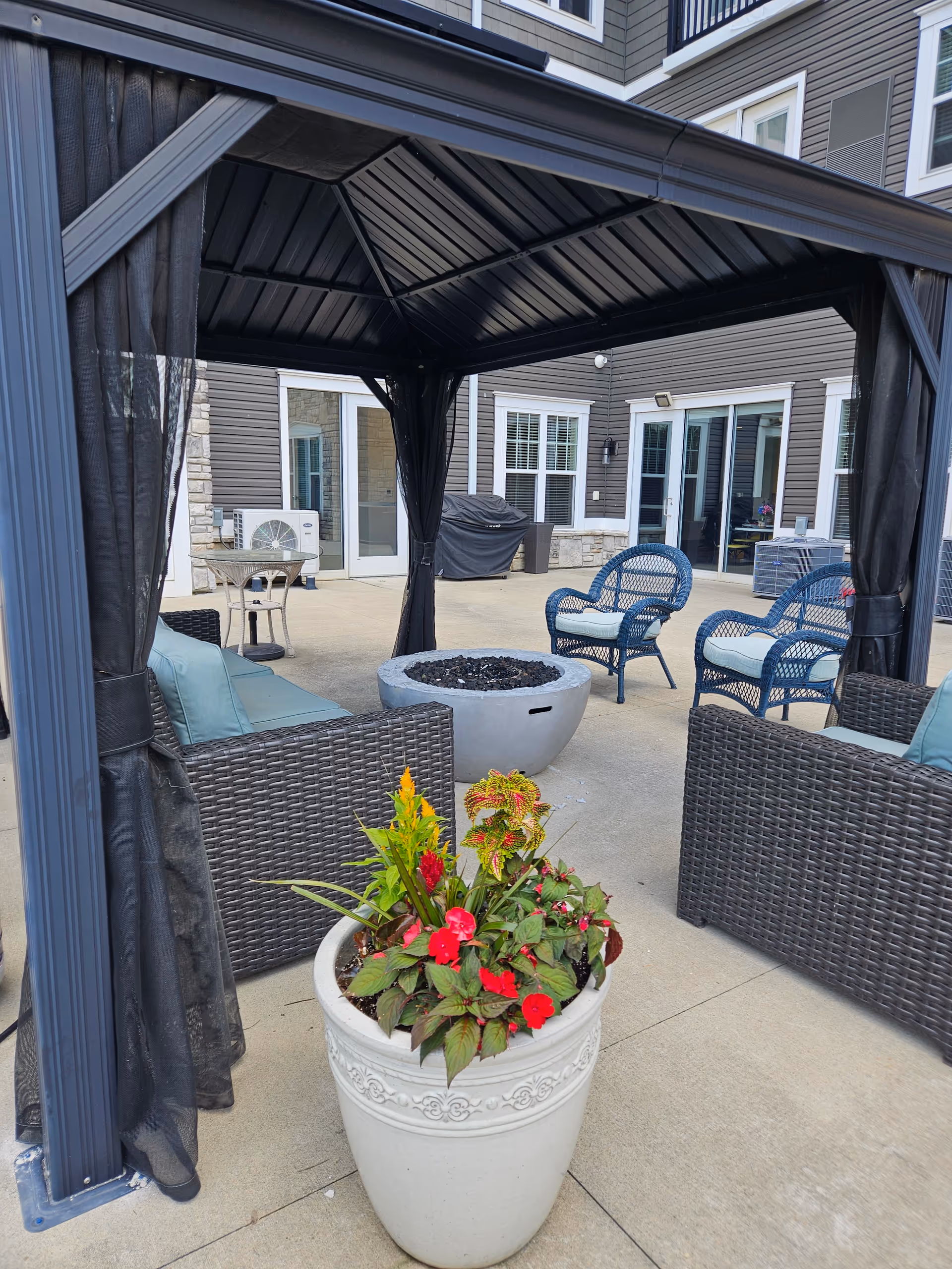 Outdoor patio area at The Echelon of Medina featuring a black metal gazebo with sheer black curtains, wicker seating with cushions, a round fire pit, and a large white planter with colorful flowers. The background shows the exterior of the building with windows and doors.