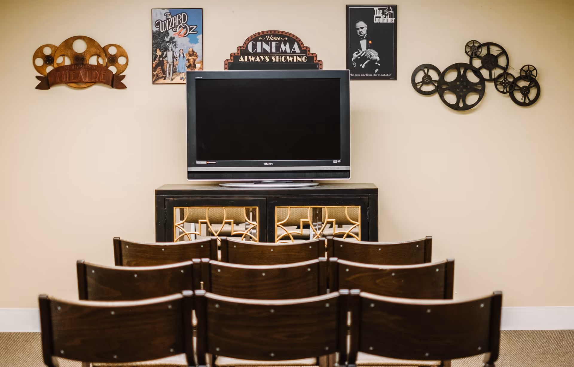 Rows of wooden chairs face a TV on a cabinet beneath movie-themed wall decorations in a small screening room.