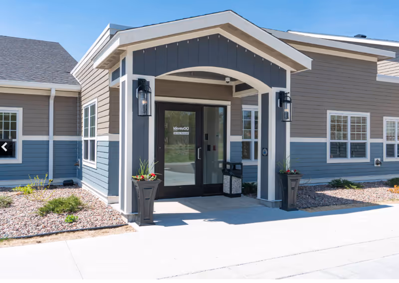 Front entrance of a senior living building with a covered portico, double glass doors, and planters.