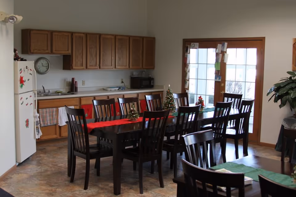 A dining area with a long dark wooden table surrounded by multiple matching chairs. The table is decorated with small Christmas trees and a red table runner. In the background, there is a kitchen area with wooden cabinets, a white refrigerator adorned with holiday magnets, a sink, and a microwave. French doors with glass panes lead outside, and a large green plant is visible on the right side of the image.