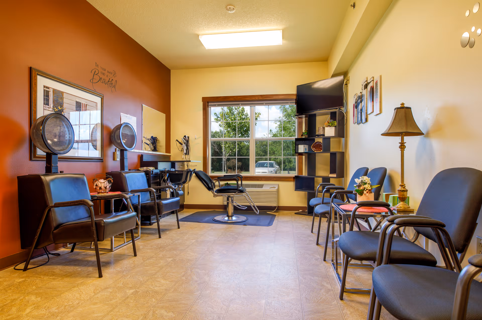 Interior view of a hair salon area in an assisted living facility. The room features a brown accent wall with two hair drying chairs and hair dryers mounted on the wall. There is a large window letting in natural light, a styling chair in front of a mirror, and several blue waiting chairs along the opposite wall. A floor lamp and a shelving unit with a TV and decorative items are also visible.