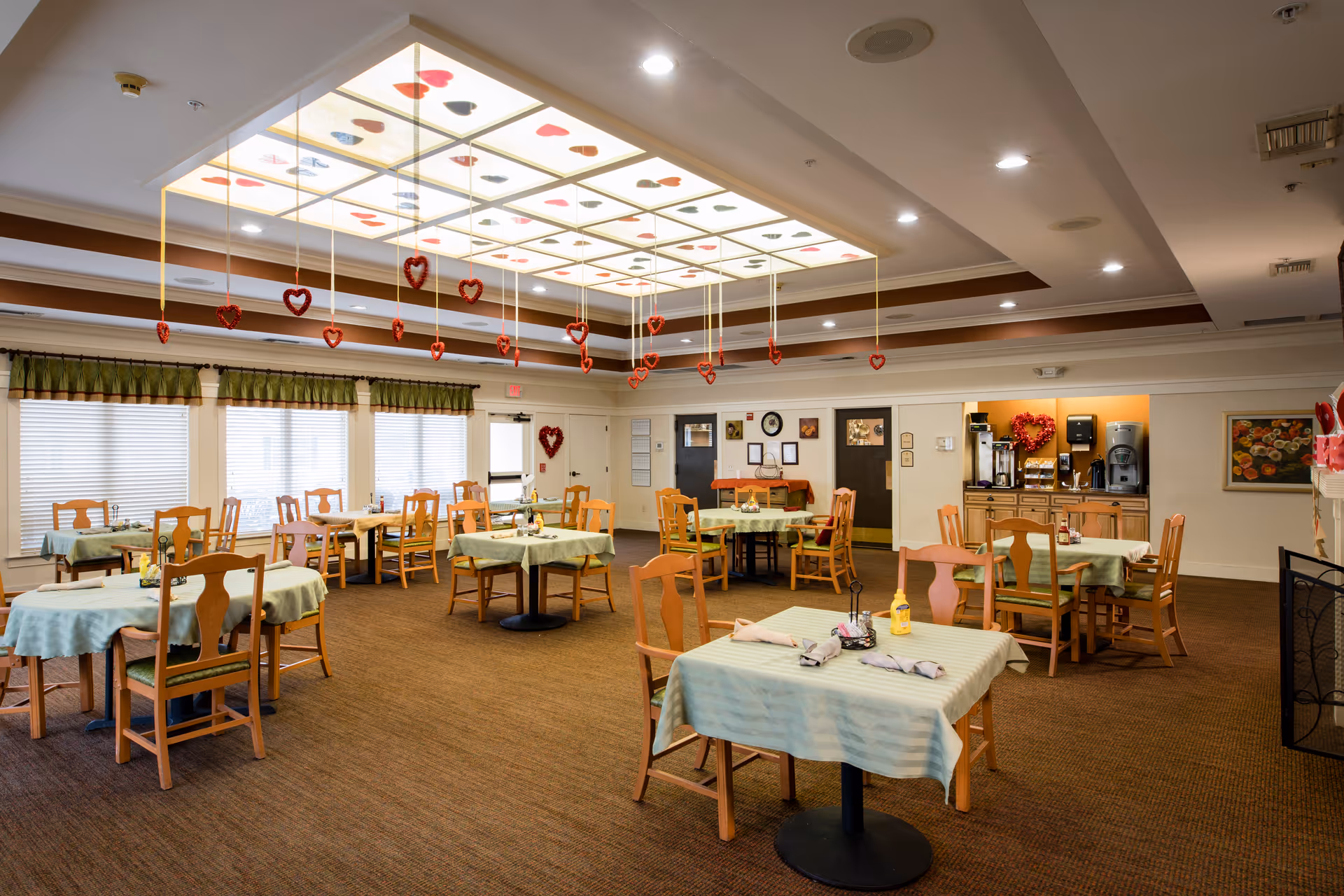 Bright, spacious dining room with multiple small tables, wooden chairs, and hanging heart decorations under a lit ceiling panel.