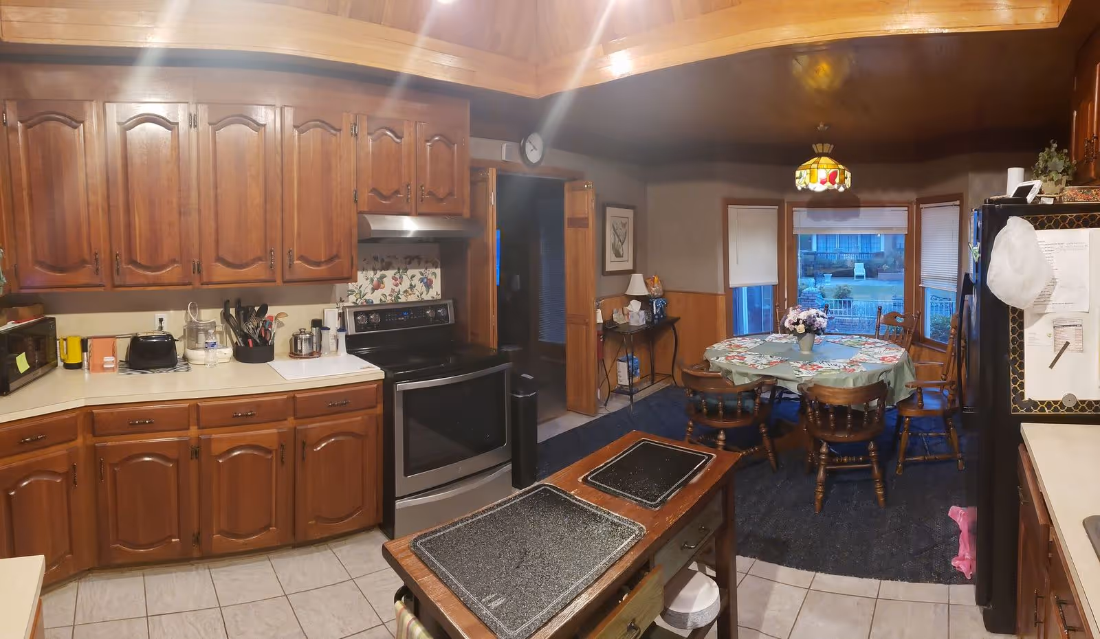 A cozy kitchen and dining area with wooden cabinets, a stainless steel stove, a small kitchen island with cutting boards, and a round dining table covered with a floral tablecloth and surrounded by wooden chairs. A stained glass pendant light hangs above the dining table, and windows with blinds look out to an outdoor area.