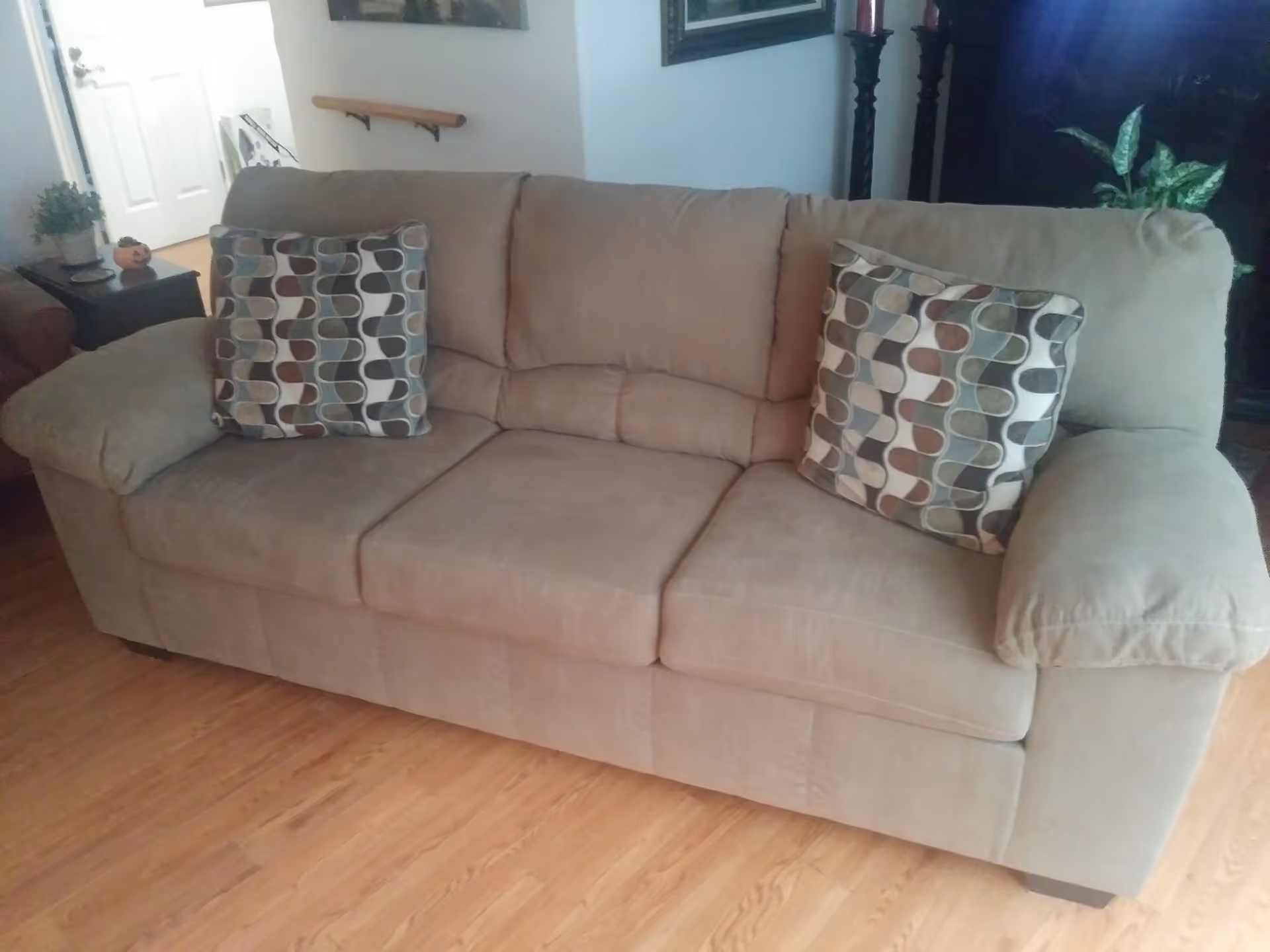 A beige fabric sofa with three seat cushions and two patterned throw pillows is placed on a wooden floor in a living room. In the background, there is a small side table with a plant and a decorative pumpkin, a white door, a handrail on the wall, and some framed artwork.