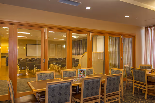 Interior view of a senior living facility dining area with wooden tables and cushioned chairs. Behind a glass partition, there is a room set up with rows of chairs, possibly for meetings or presentations. The room is well-lit with ceiling lights and has curtains on the windows.