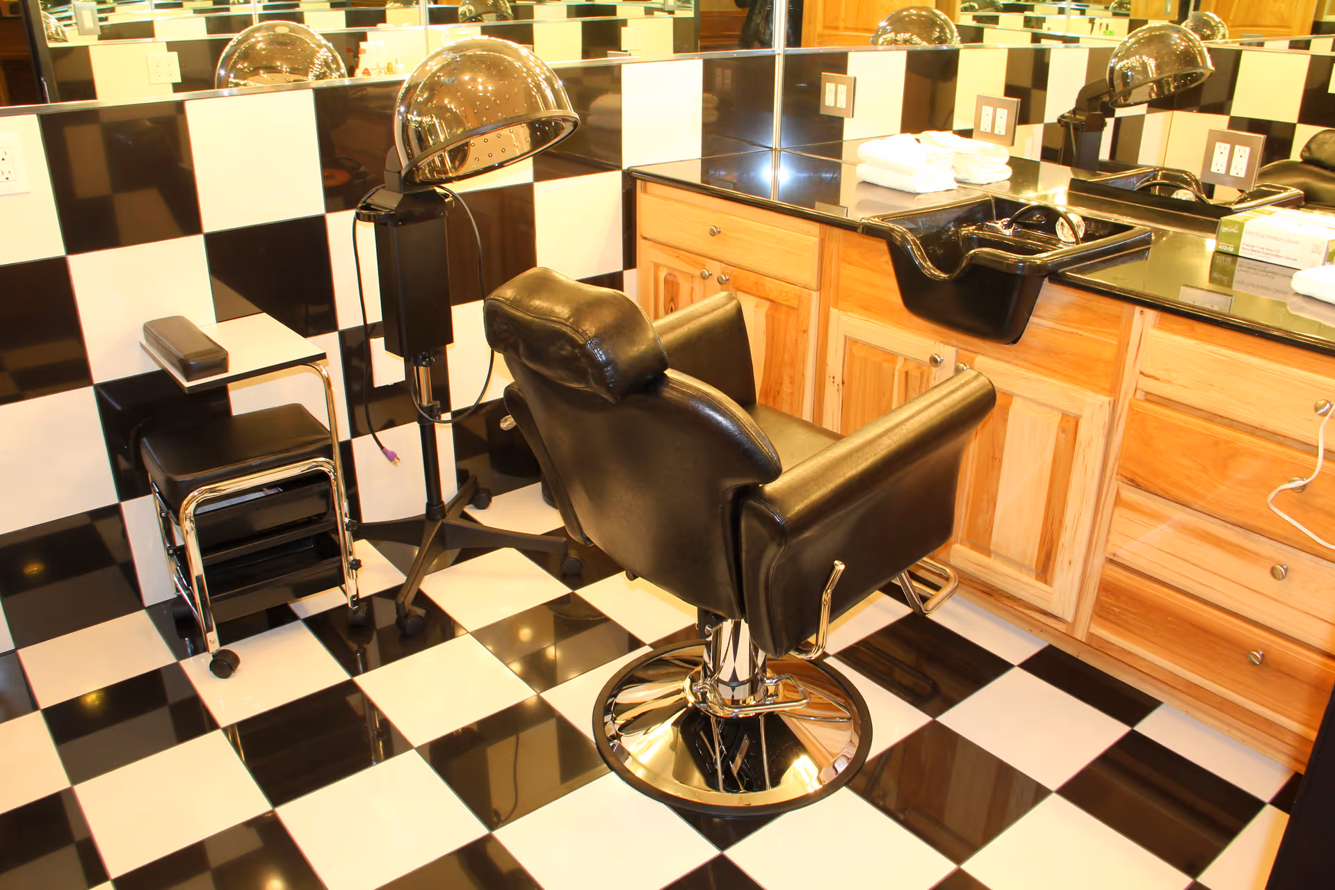 A black salon chair and hooded hair dryer next to a wash basin and wooden cabinets on a black-and-white checkered floor.