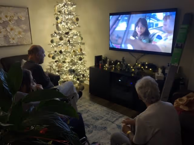 Three elderly people sitting in a dimly lit living room watching a movie on a wall-mounted TV. A decorated and lit Christmas tree is visible in the corner, along with a dark cabinet beneath the TV adorned with holiday decorations.