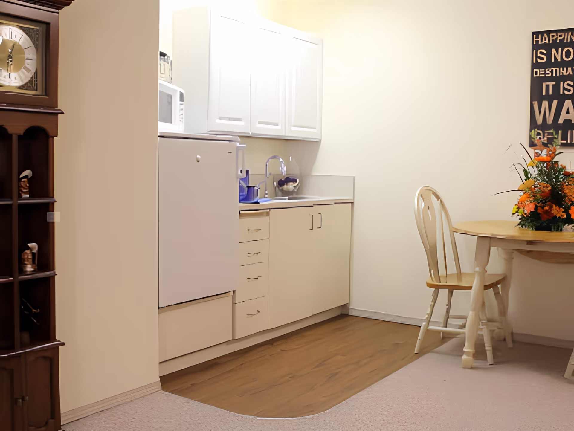 A small kitchen area with white cabinets, a white refrigerator, a microwave, and a sink. To the right, there is a wooden dining table with a chair and a floral centerpiece. A decorative clock is partially visible on the left side of the image, and a wall sign with partially visible text is on the right.