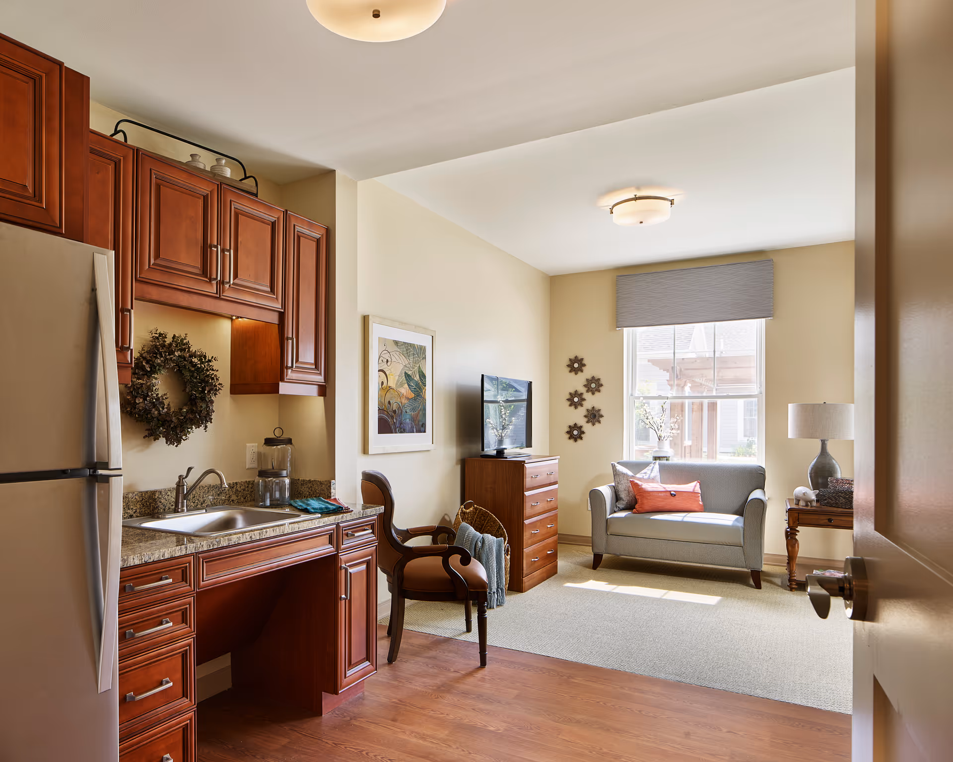 A cozy interior space featuring a small kitchen area with wooden cabinets, a stainless steel refrigerator, and a sink. Adjacent to the kitchen is a living area with a cushioned loveseat, a wooden chest of drawers with a TV on top, a wooden chair, and a side table with a lamp. The room is well-lit by natural light coming through a window with a gray shade.