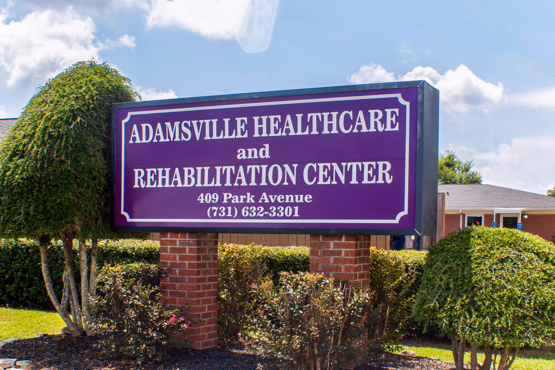 Purple roadside sign reading "Adamsville Healthcare and Rehabilitation Center" in front of trimmed bushes and the facility building.