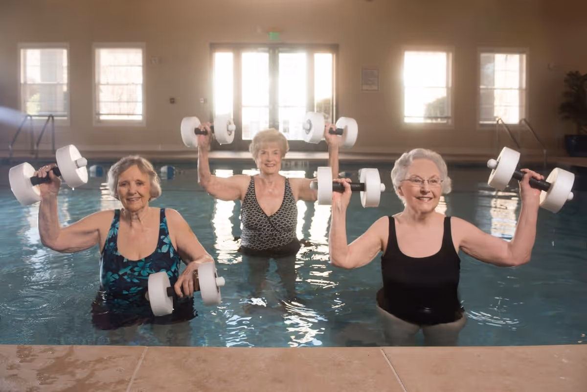 Three elderly women standing in an indoor swimming pool, each holding white water dumbbells above the water, smiling and exercising together.