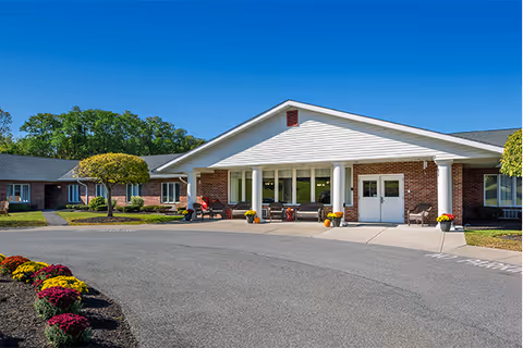 Front exterior view of a single-story senior living facility building with a covered entrance supported by white columns. There are chairs and potted plants near the entrance, a curved driveway, and well-maintained landscaping including trimmed bushes and colorful flowers. The sky is clear and blue.