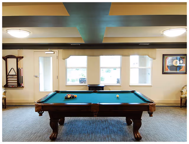 A well-lit room with a pool table in the center, set up for a game with balls arranged in a triangle and a cue ball placed separately. The room has three large windows letting in natural light, a door on the left side, a wall-mounted pool cue rack, and framed artwork featuring an 8-ball on the right wall. Two chairs are partially visible on either side of the room.