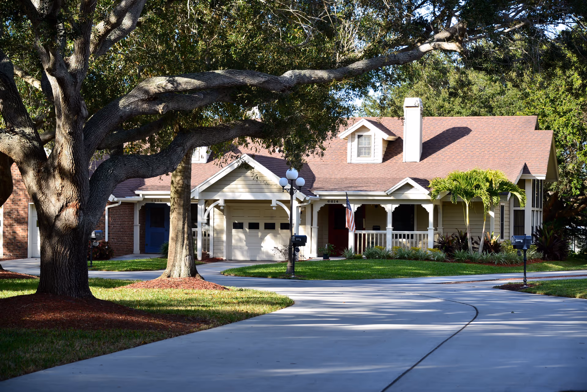 A single-story residential building with a beige exterior, brown shingled roof, and a small front porch with white railings. The house has a garage with a closed door and an American flag hanging near the entrance. There are large trees and well-maintained green lawns surrounding the house, with a curved concrete driveway leading up to it.