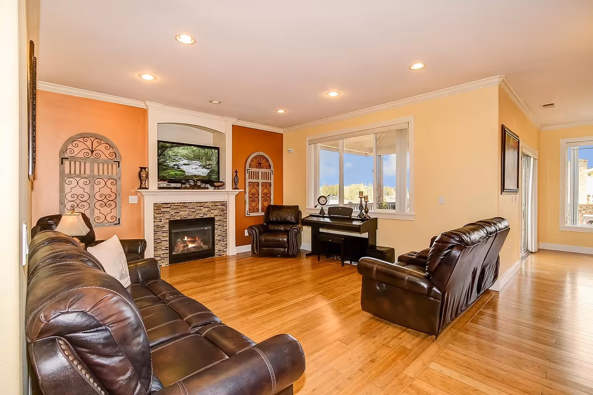 A bright living room with wooden flooring, featuring a dark brown leather sofa and two matching armchairs. There is a fireplace with a mosaic tile surround and a TV mounted above it. The walls are painted in warm tones of orange and beige, with decorative wrought iron wall art. A piano is placed near a large window that lets in natural light and offers a view of the outdoors.